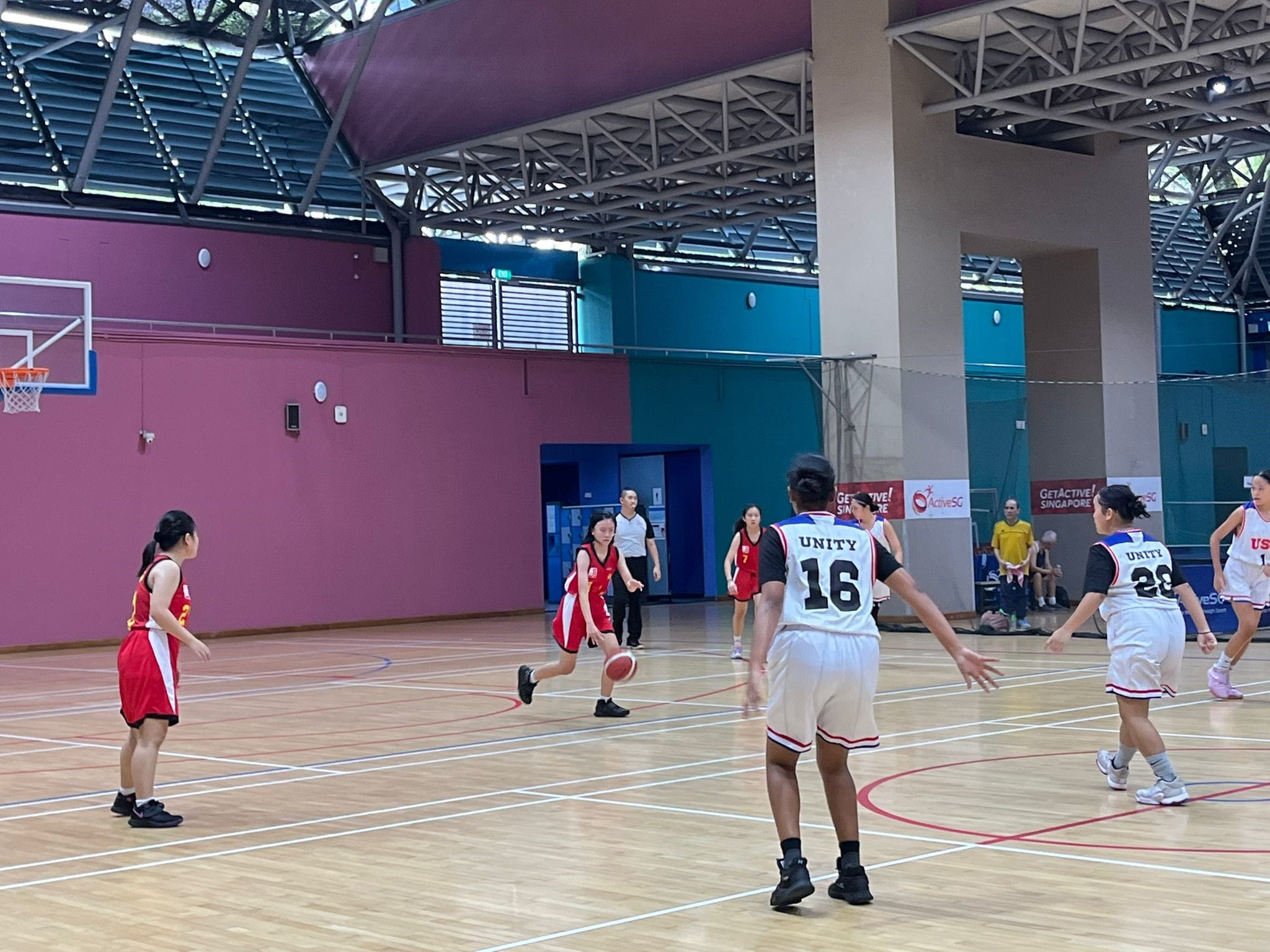 Girls in red and white Unity jerseys play basketball on an indoor court with pink and teal walls.